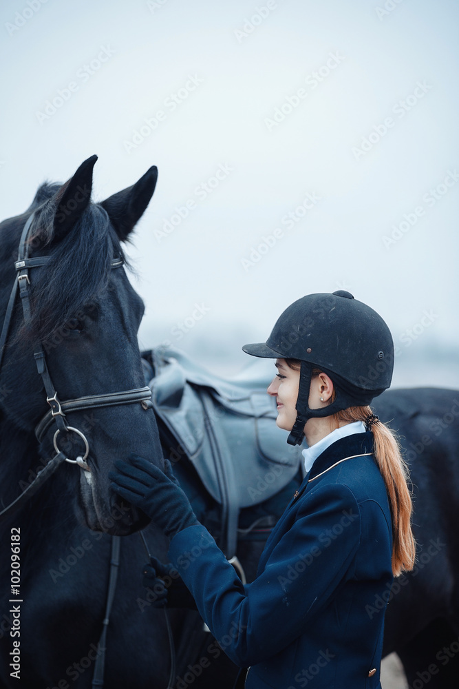 a tall girl with long blond hair in a jockey outfit with a beautiful black horse in an empty snow-covered field in winter a tall girl with long blond hair in a jockey outfit with a beautiful black horse in an empty snow-covered field in winter