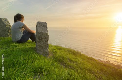 Mujer joven observa el atardecer sobre el mar desde lo alto de un acantilado, sentada sobre el césped photo
