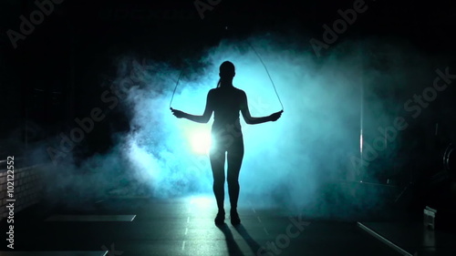 Sporty woman jumps on the jumping rope in silhouette and smoke, front view, slow motion