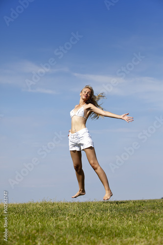The happy woman in white bikini and shorts jumps in a summer green field