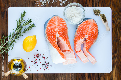 two steaks of salmon with a sprigs of rosemary, lemon, pepper,  sea salt, olive oil and vinegar in glass jar, herbs in wooden scoop on a white cutting board on wooden background, top view.