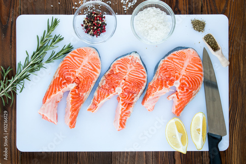 steaks of salmon with a sprigs of rosemary, lemon, pepper,  sea salt, , herbs in wooden scoop,  chef's knife on a white cutting board on wooden background, top view.