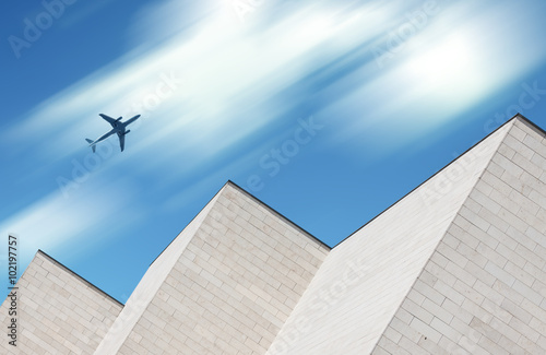 Airplane flying over modern white brick building with motion blur clouds in the background 