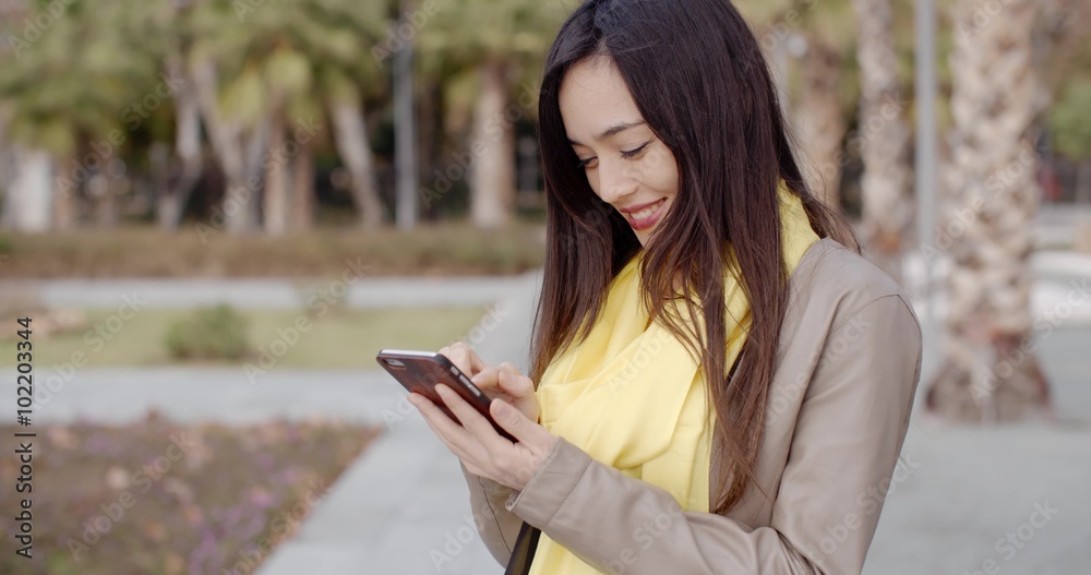 Stylish young woman checking a message on her mobile phone as she stands in an urban park close up upper body Stylish young woman checking a message on her mobile phone as she stands in an urban park close up upper body