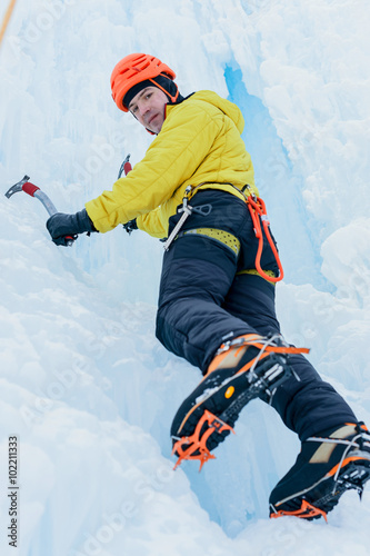Athletic alpinist man in orange helmet and ice tools axe climbing a large wall of ice. Outdoor Sports Portrait