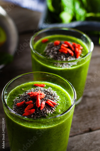 Fotografie Fresh green smoothie sprinkled with chia seeds and goji berries in the background fruits and vegetables on wooden table