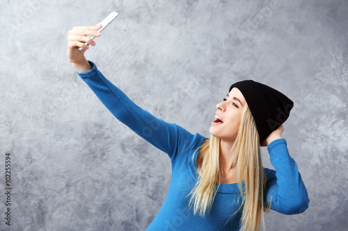 Young girl in black hat taking photo of her self with smart phone on grey wall background