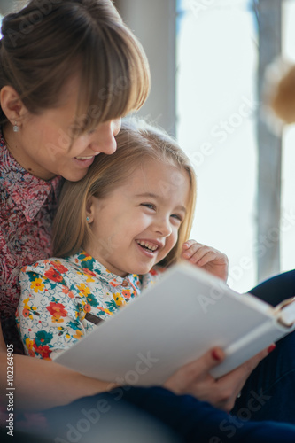Portrait of a smiling young cute mother and daughter reading a book lying and relax on the bed in a bright big white room 