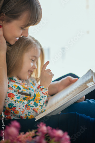 Portrait of a smiling young cute mother and daughter reading a book lying and relax on the bed in a bright big white room 