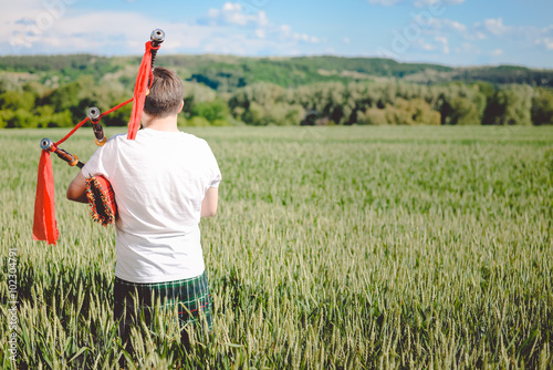 Back view of man enjoying playing pipes in Scotish traditional kilt on green outdoors summer field. photo