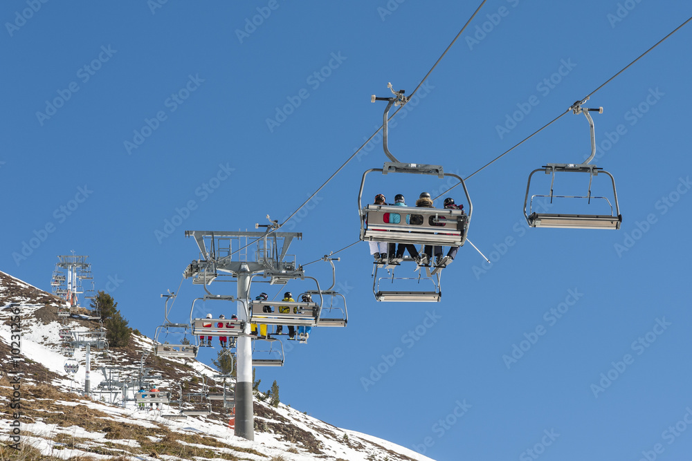 Skiers sitting on a chairlift at a ski resort, with snowy mountains on background.