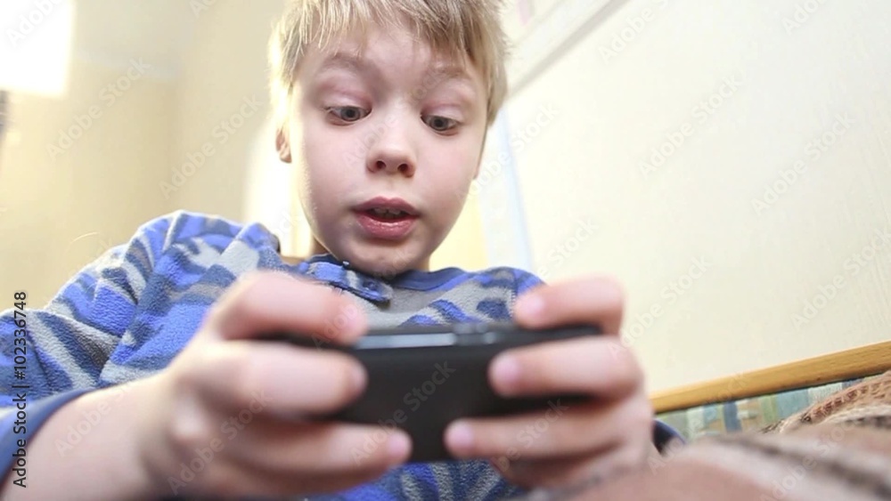 Funny 8 years old kid playing computer games laying in his bed in morning. Child using smartphone. Cute blond caucasian boy dressed in blue pajamas. Emotional beautiful child portrait. Focus on face Funny 8 years old kid playing computer games laying in his bed in morning. Child using smartphone. Cute blond caucasian boy dressed in blue pajamas. Emotional beautiful child portrait. Focus on face