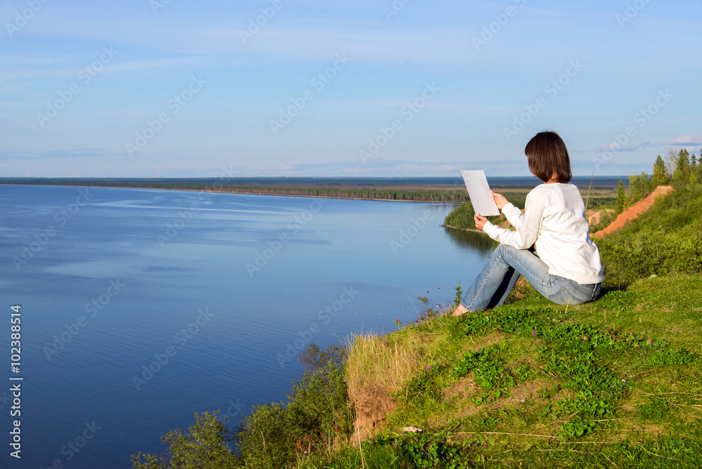 Young woman sitting on the shore of the lake with blank sheet of