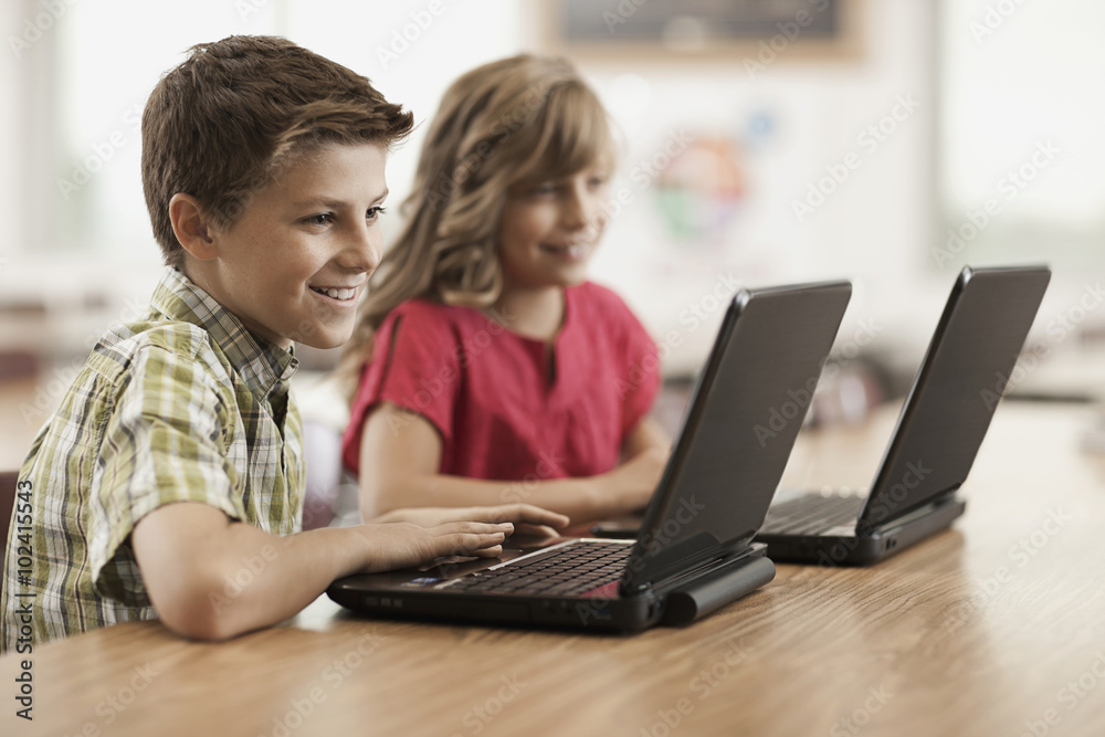 Two children seated at desks in class using laptop computers.