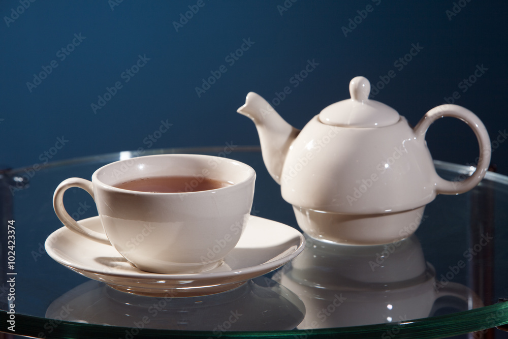 White ceramic cup of tea with saucer and teapot on a glass table with a blue background