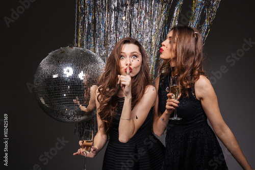 Two amusing women showing silence gesture and drinking champagne
