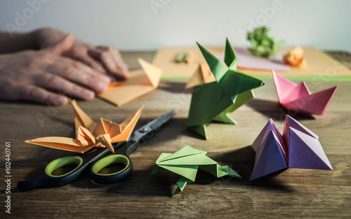 Origami figures, scissors and pencils on wooden table, in the background hands folding colored paper photo