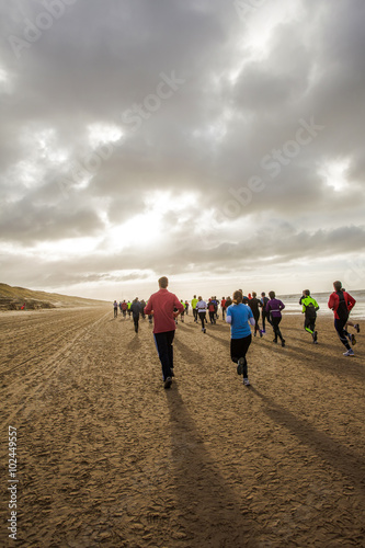 A group of back lit people in colorful outfits are running on a beach during the half marathon of Egmond. 