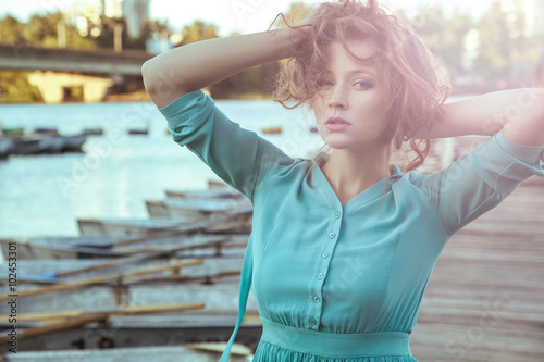 Beautiful young fashion model in green turquoise dress and fashion makeup and hairstyle is posing on pier, posing and looking at camera. close up. sunset effect on camera. with passion.