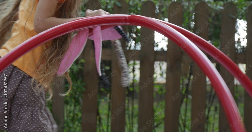 Girl is Swinging on The Carousel Holding Pink Toy Tilda Rabbit Little Girl With Long Fair Hairs in Orange T-Shirt is Playing at Playground Wooden Fence Girl is Swinging on The Carousel Holding Pink Toy Tilda Rabbit Little Girl With Long Fair Hairs in Orange T-Shirt is Playing at Playground Wooden Fence
