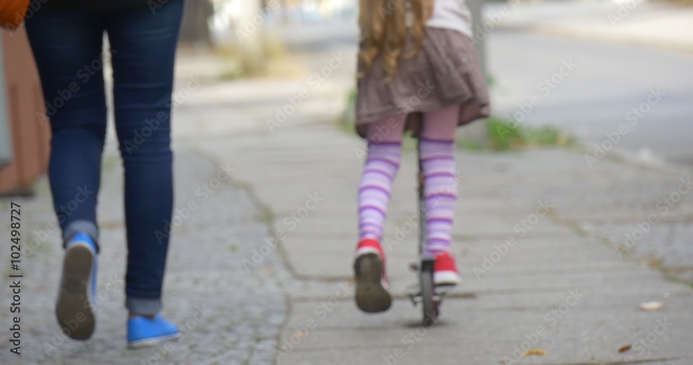 Little Blonde Girl is Riding The Kick Scooter by Street Red Sneakers Close Up Tilt Up Woman Mom is Walking along Girl Daughter People are Walking Away Little Blonde Girl is Riding The Kick Scooter by Street Red Sneakers Close Up Tilt Up Woman Mom is Walking along Girl Daughter People are Walking Away