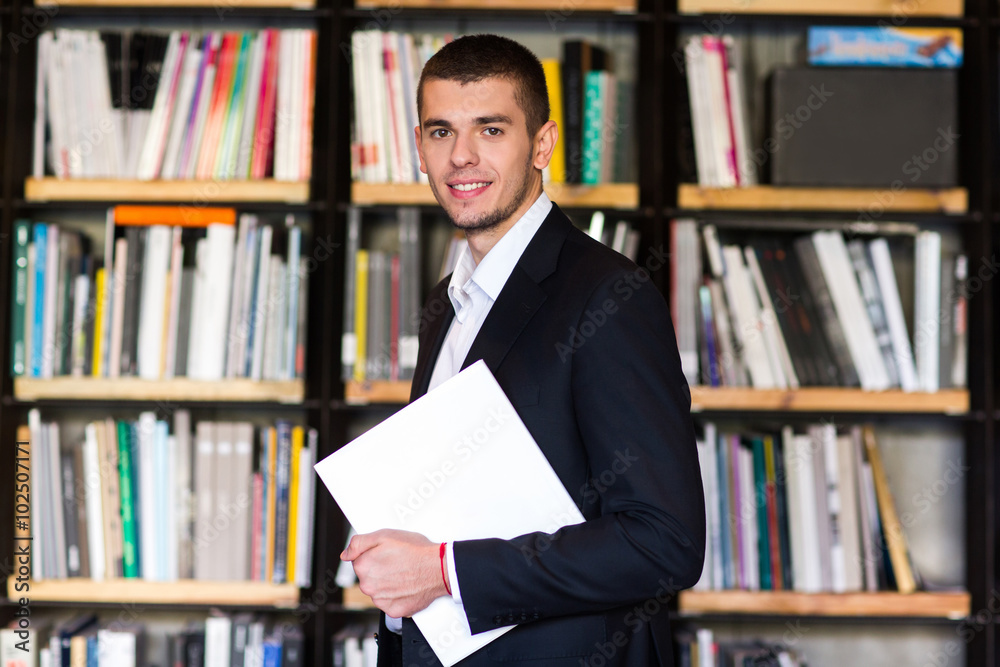 Student in library. Handsome young man holding books and smiling while standing in library Student in library. Handsome young man holding books and smiling while standing in library