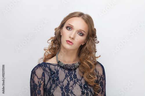 Young curious blonde teen model in blue dress with curly hairstyle posing on white studio background