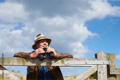 Senior Man With Binoculars Leaning Against Wooden Gate photo