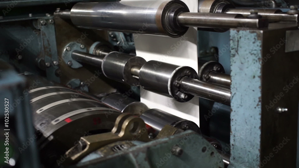 Handheld shot of slow moving newsprint or newspaper being fed through rollers at the end of an industrial scale newspaper printing offset press with the paper cutting drum in the foreground.