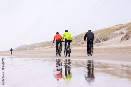 four unrecognizable people drive with mountain bikes on a beach during a contest. with a visible reflection in the water in the foreground. 
