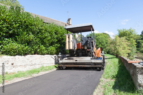 Road tarring machine using a premix asphalt in a village tarring a narrow rural road viewed from behind showing the newly compacted tarmac
