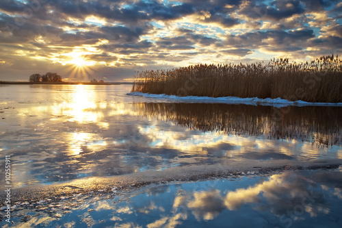 Winter landscape with river, reeds and cloudy sunset sky. 