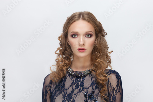 Closeup portrait of young adorable blonde woman in blue dress with cute makeup and wavy hairstyle questioningly looking into camera posing on white studio background
