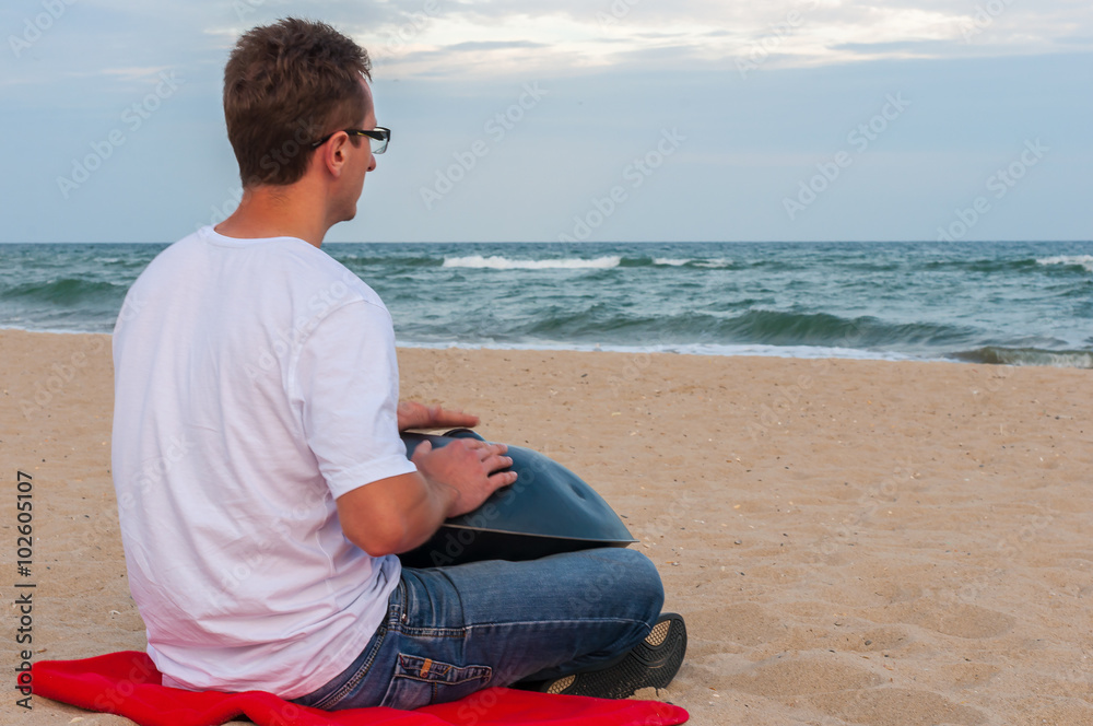 Young stylish guy sitting on the sand beach and playing handpan or hang  with sea On Background. The Hang is traditional ethnic drum musical  instrument Stock Photo | Adobe Stock
