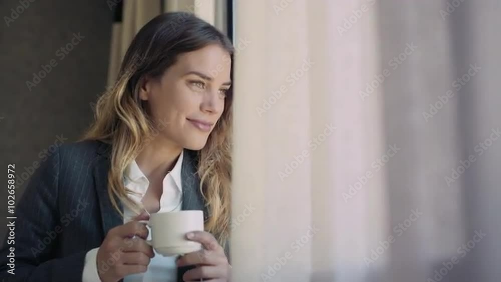 Young business woman at her hotel bedroom drinking a cup of coffee and looking through the window smiling. Close up. Young business woman at her hotel bedroom drinking a cup of coffee and looking through the window smiling. Close up.