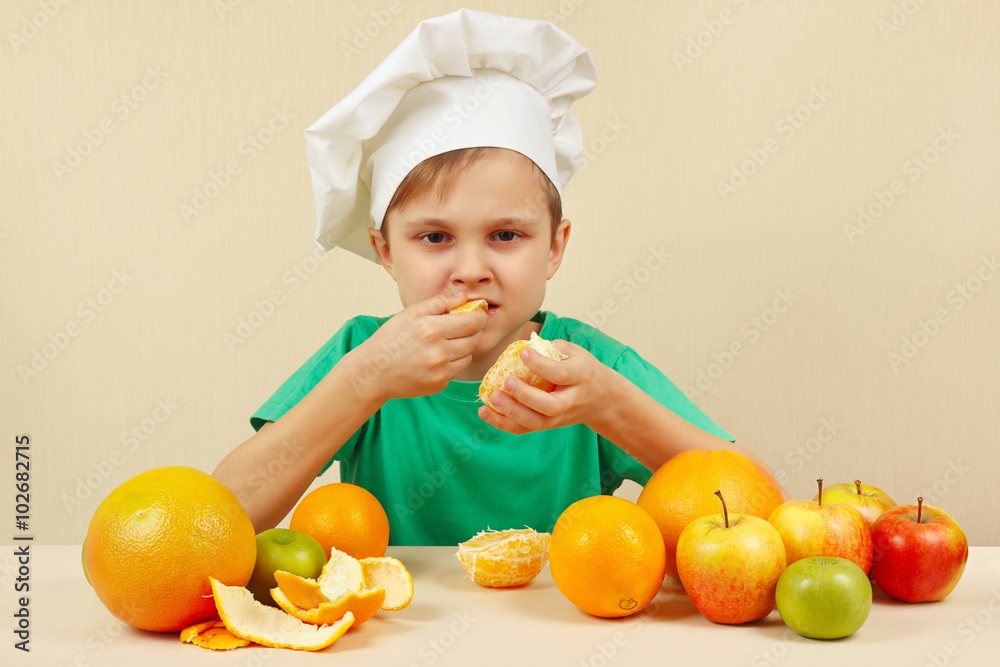 Little funny boy eat fresh orange at the table with fruits Little funny boy eat fresh orange at the table with fruits