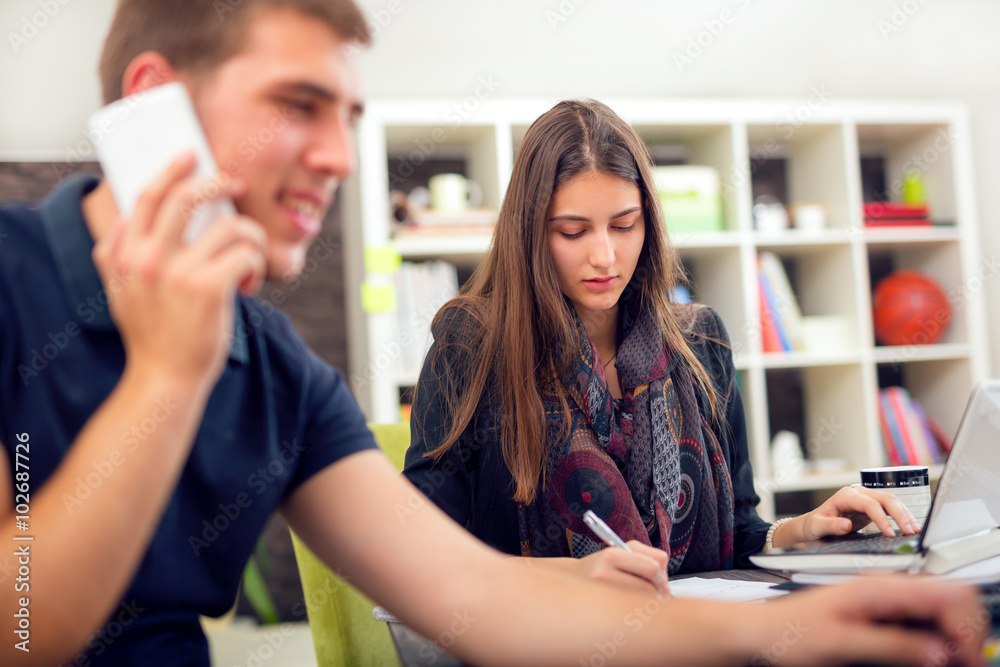 Beautiful smiling young  businesswoman sitting working with a young man in a busy office.  Man using a smart phone