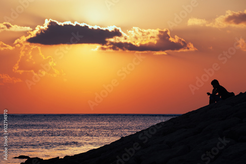 Black silhouette of thinking man sitting alone on the sea beach reading message on mobile phone on sunset sky background. Lifestyle, single people emotions, family conflicts and problem concepts.