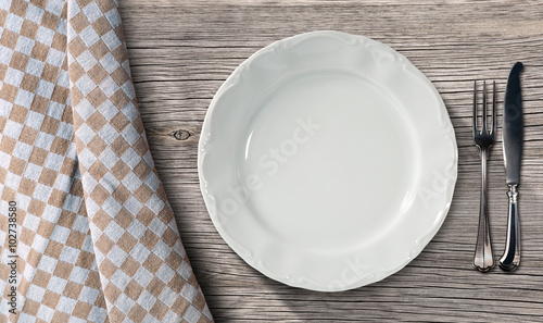 Plate and Cutlery on a Table with Tablecloth / Empty white plate on a table in pine wood with silver cutlery, fork and knife, and a brown and white checkered tablecloth 