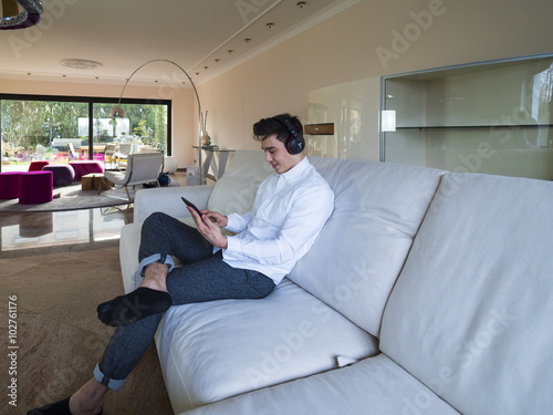 Ein junger Mann , etwa 20 Jahre, sitzt im Wohnzimmer  auf einem Sofa mit Kopfhöhrern und schaut in seinen Laptop