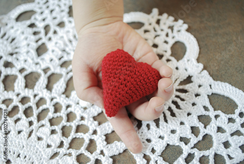 Child's hand holding crochet red heart on white napkin background.