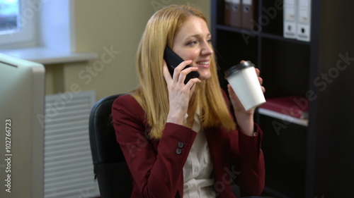 Coffee-break. Businesswoman talking on his mobile phone, spinning on a chair and drinking coffee in the office.