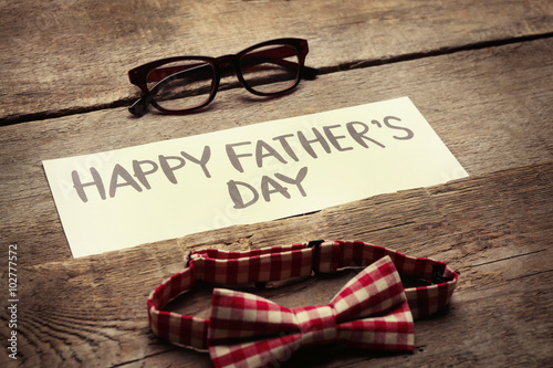 Happy Father's Day inscription with red bow tie and glasses on wooden background. Greetings and presents