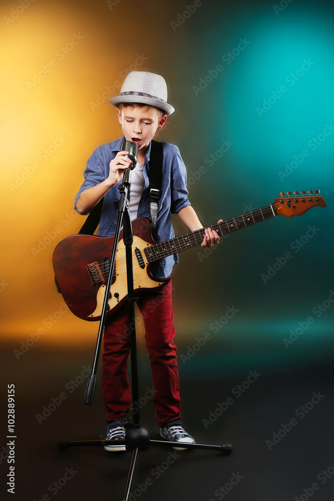 Naklejka premium Little boy playing guitar and singing with microphone on a dark lighted background