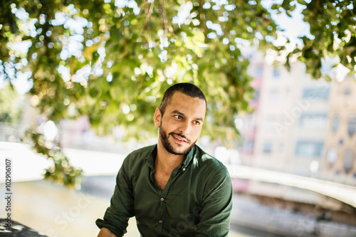 Young bearded man sitting outdoors under trees and smiling