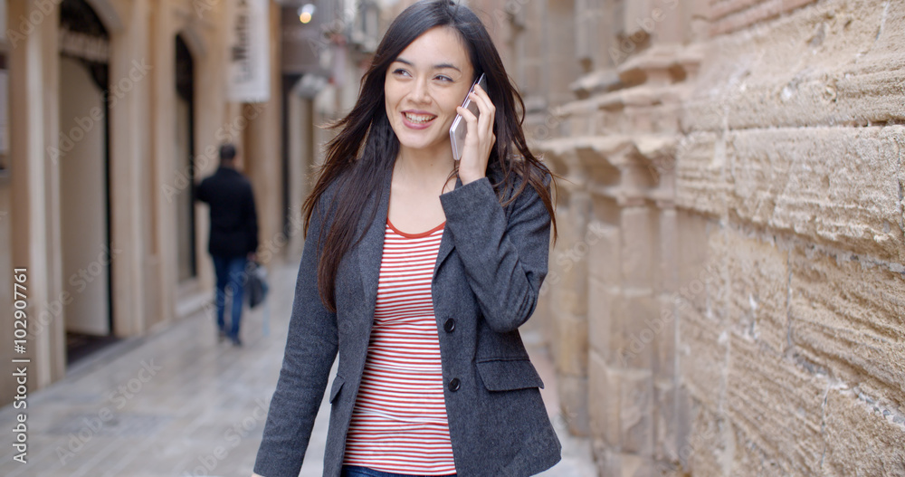 Young woman walking through town with her mobile phone listening to a call with a smile as she strides along a narrow alley