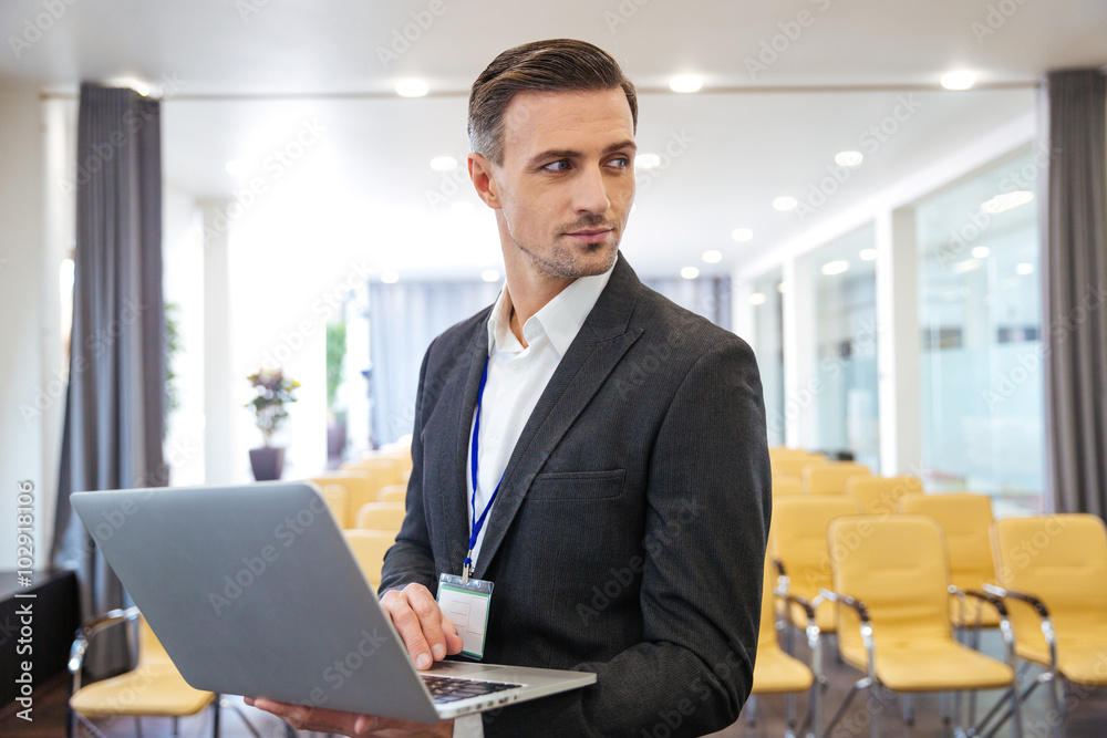 Serious businessman holding laptop in empty meeting hall Serious businessman holding laptop in empty meeting hall