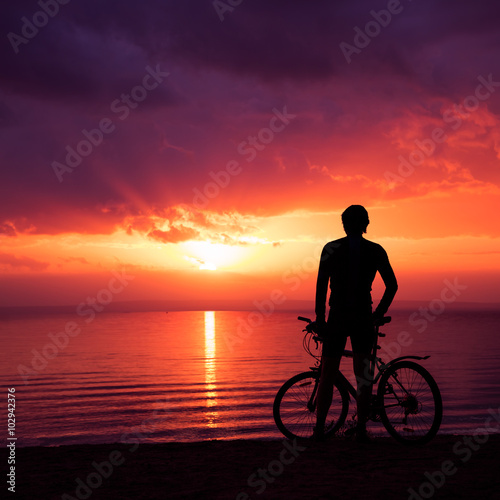 Man Standing with a Bike at Sunset by the Sea © Maryia Bahutskaya