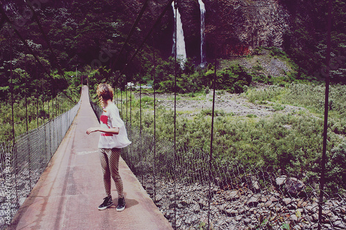 girl in t-shirt standing on bridge and looking back at waterfall