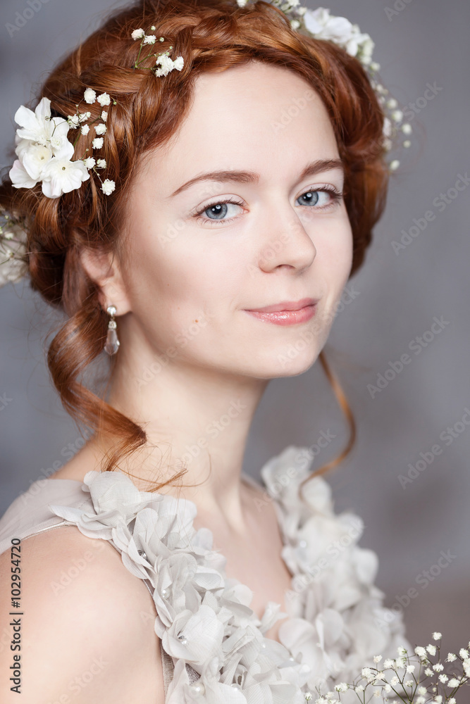 Portrait of beautiful red-haired bride. She has a perfect pale skin with delicate blush. White flowers in her hair. She smiles gently. She has light gray eyes.